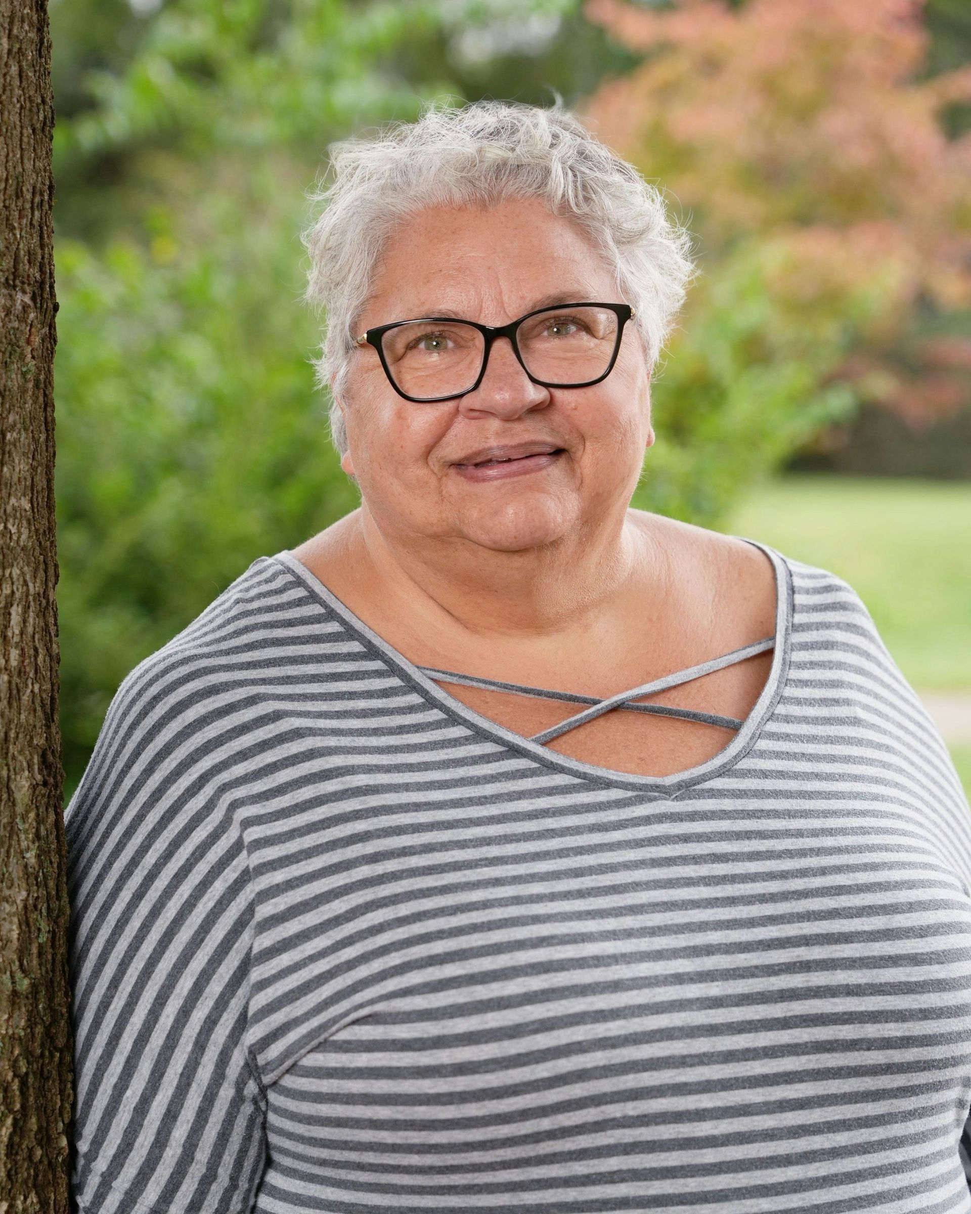 Woman with gray hair and glasses, wearing a striped shirt, leaning against a tree outdoors.
