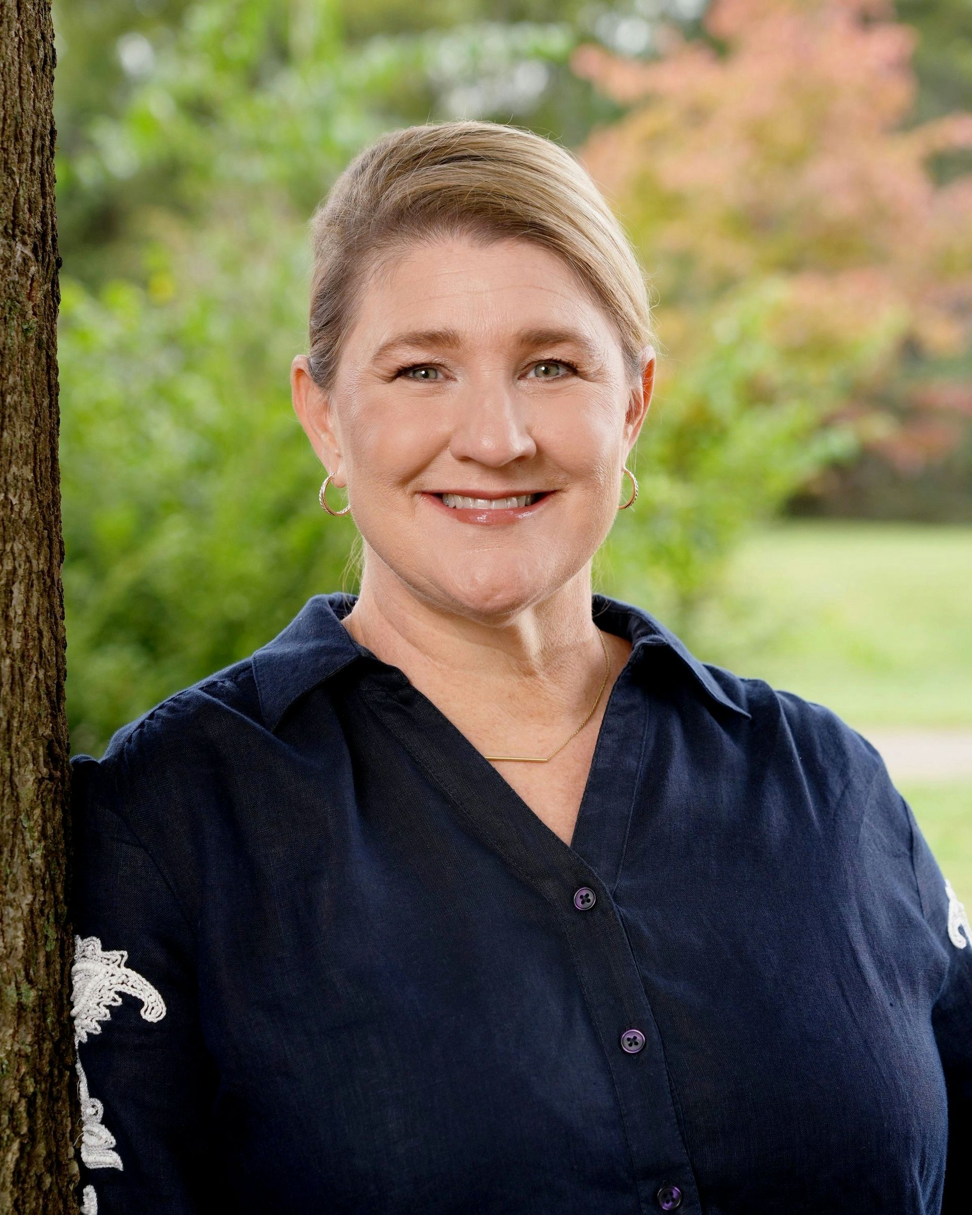 Woman with blond hair, smiles, wearing a navy shirt, leaning against a tree in a park.