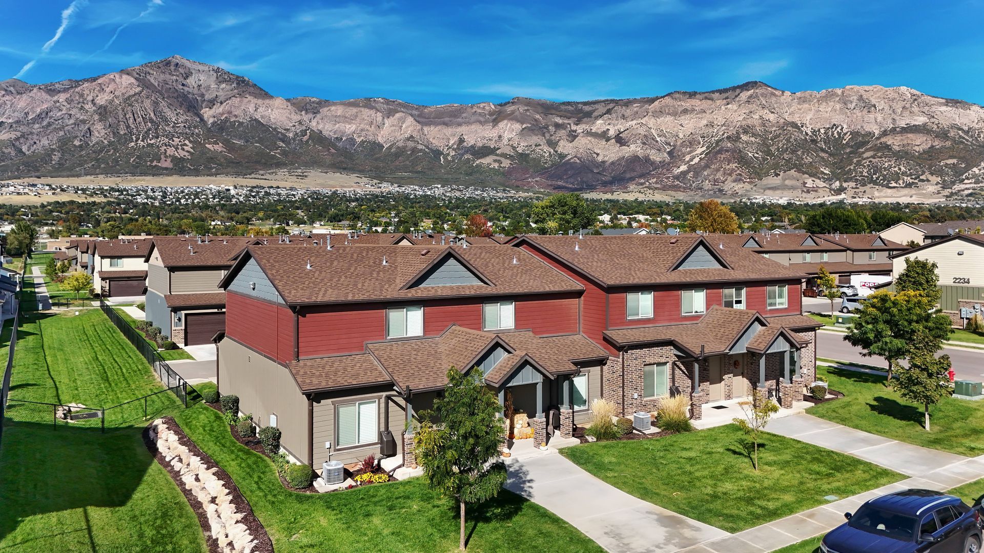 Townhouses with red and tan siding, brown roofs, green lawns, and mountains in the background.
