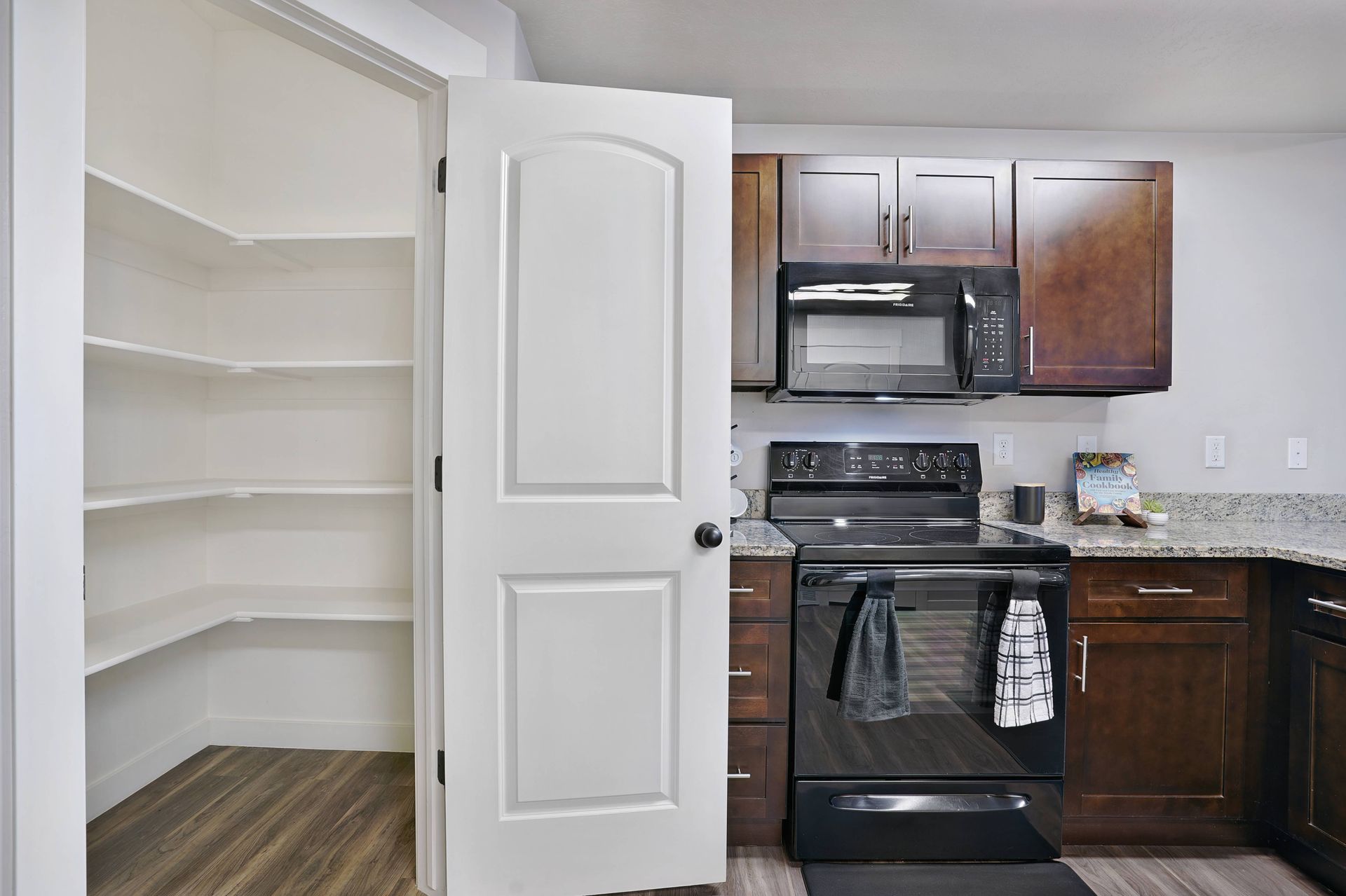 Kitchen with pantry: open white door reveals shelves, dark cabinets, appliances, and countertops.
