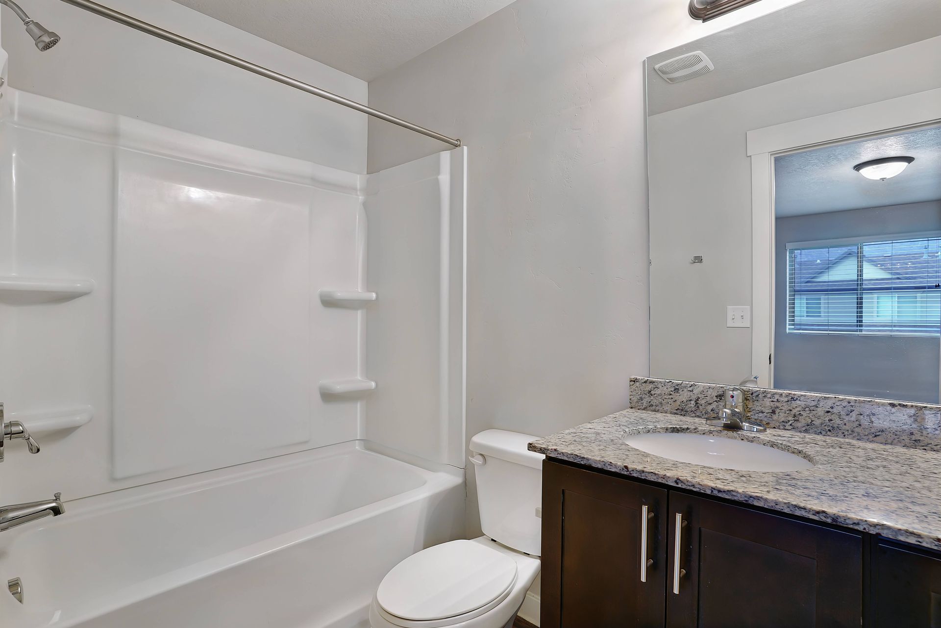 Bathroom with white tub/shower, toilet, dark vanity with granite countertop, and large mirror.