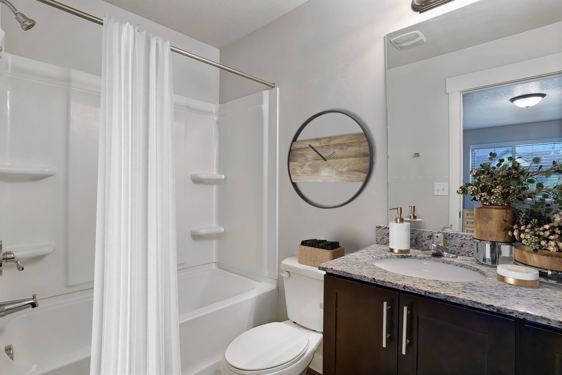 Bathroom with white tub/shower, toilet, dark vanity with a granite countertop, and a decorative clock.
