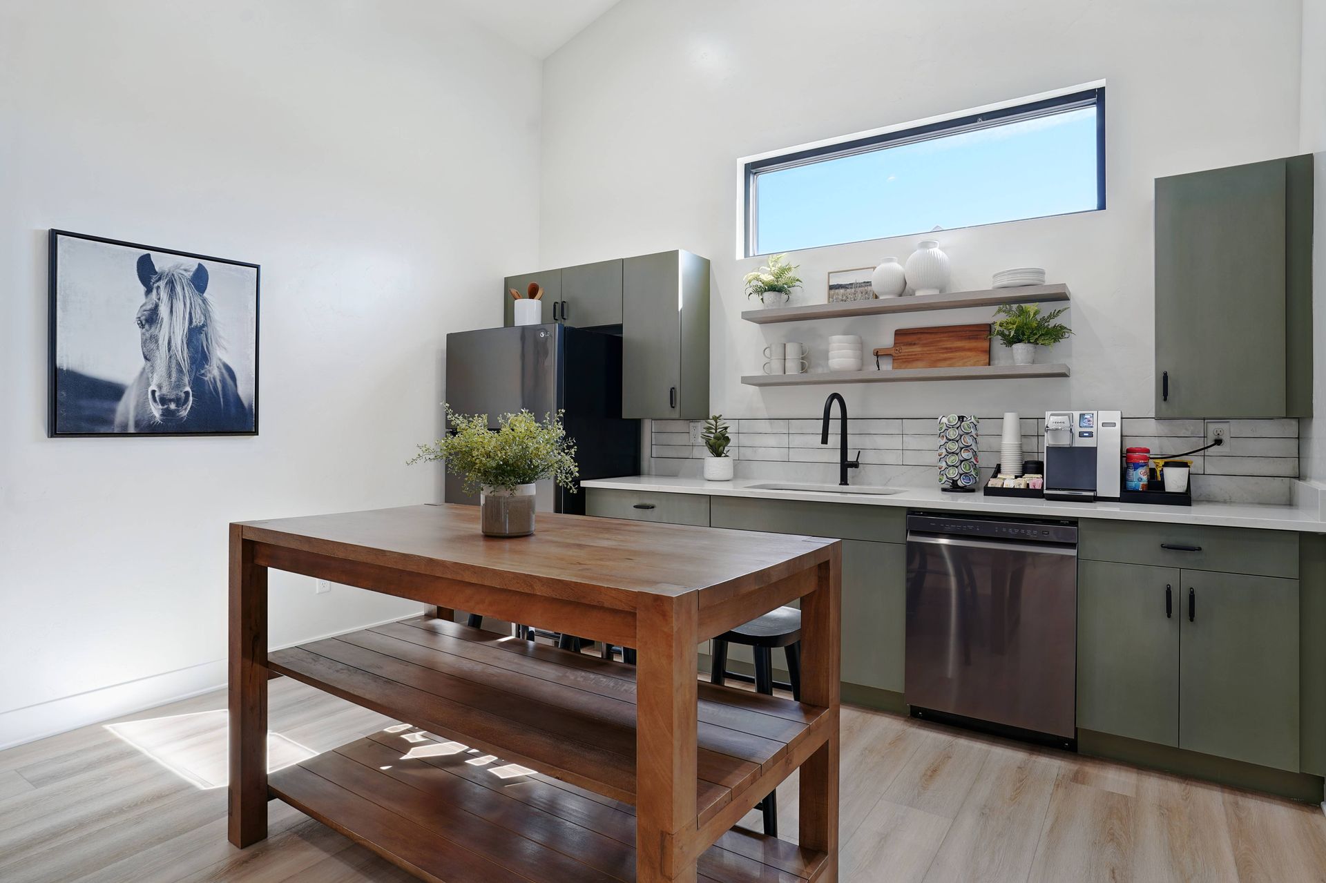 Modern kitchen with green cabinets, stainless steel appliances, and a wooden island. White walls and artwork.