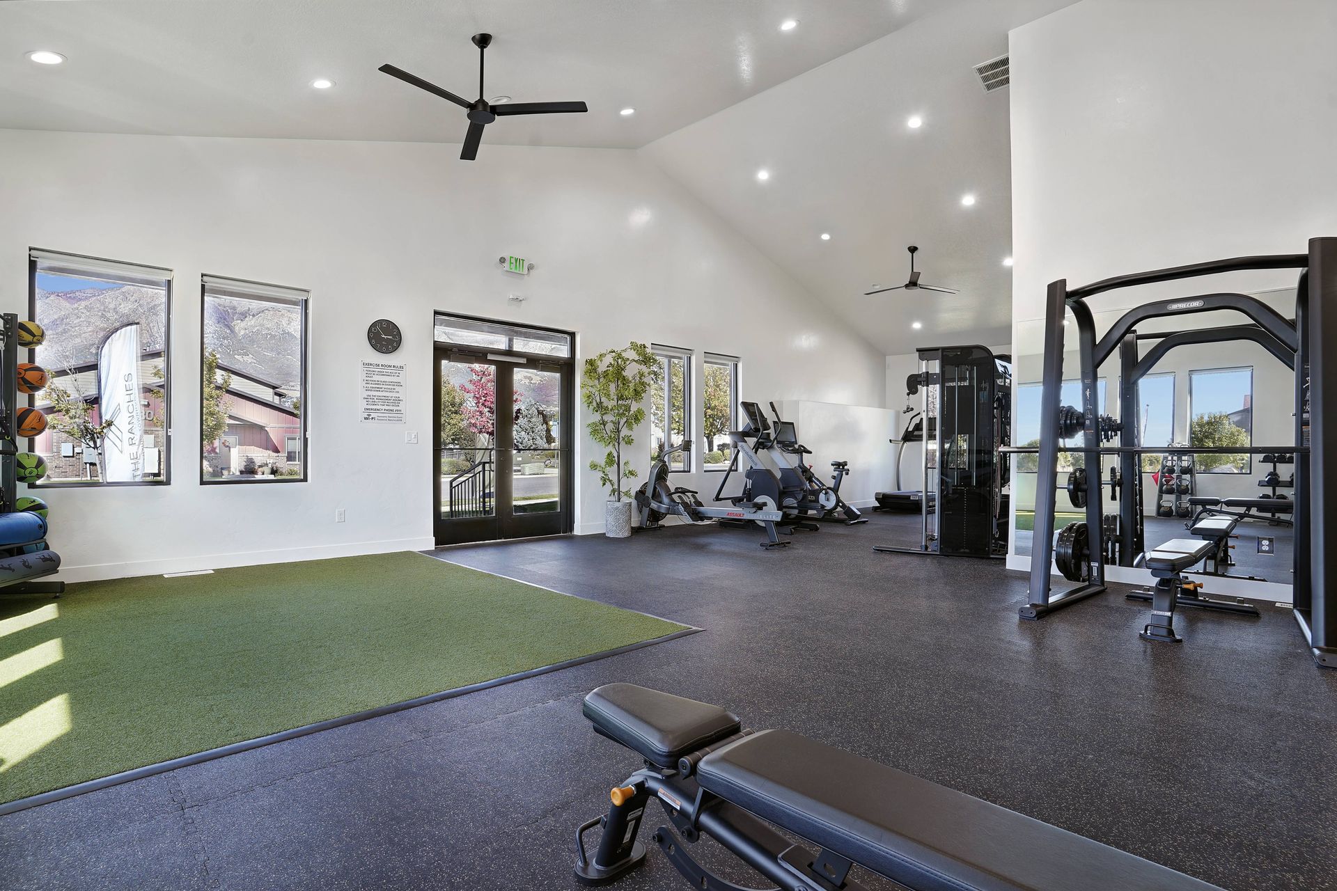 Gym interior with exercise equipment on a black floor, windows, and high ceiling. Artificial turf patch in foreground.