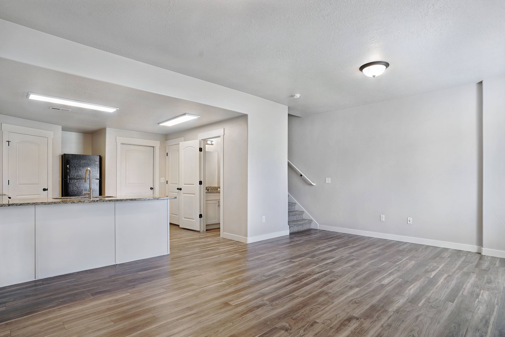 Empty modern living space with kitchen and stairs, white walls, wood flooring, and neutral colors.