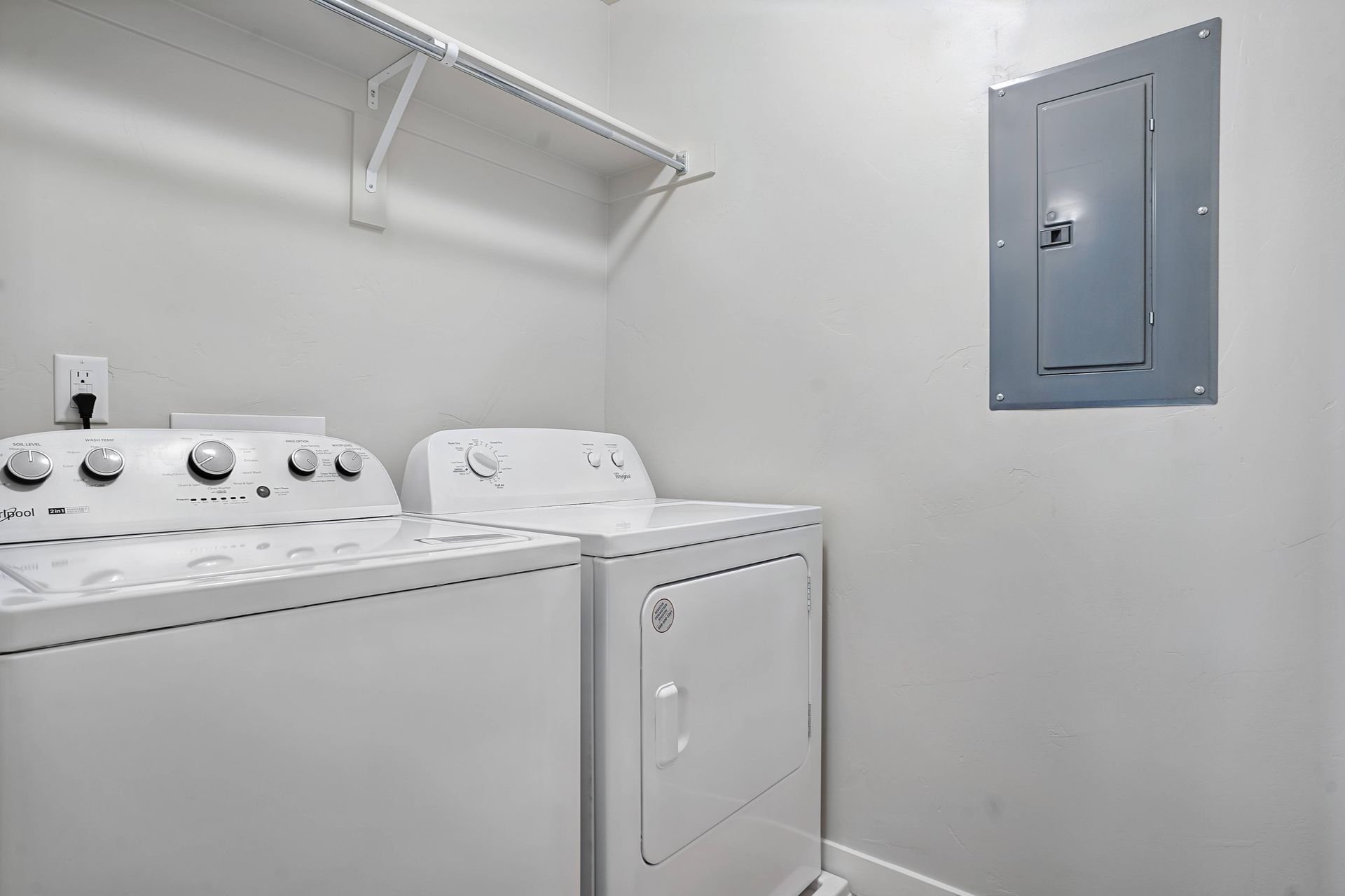 A laundry room with a white washer and dryer, shelf, and electrical panel on the wall.