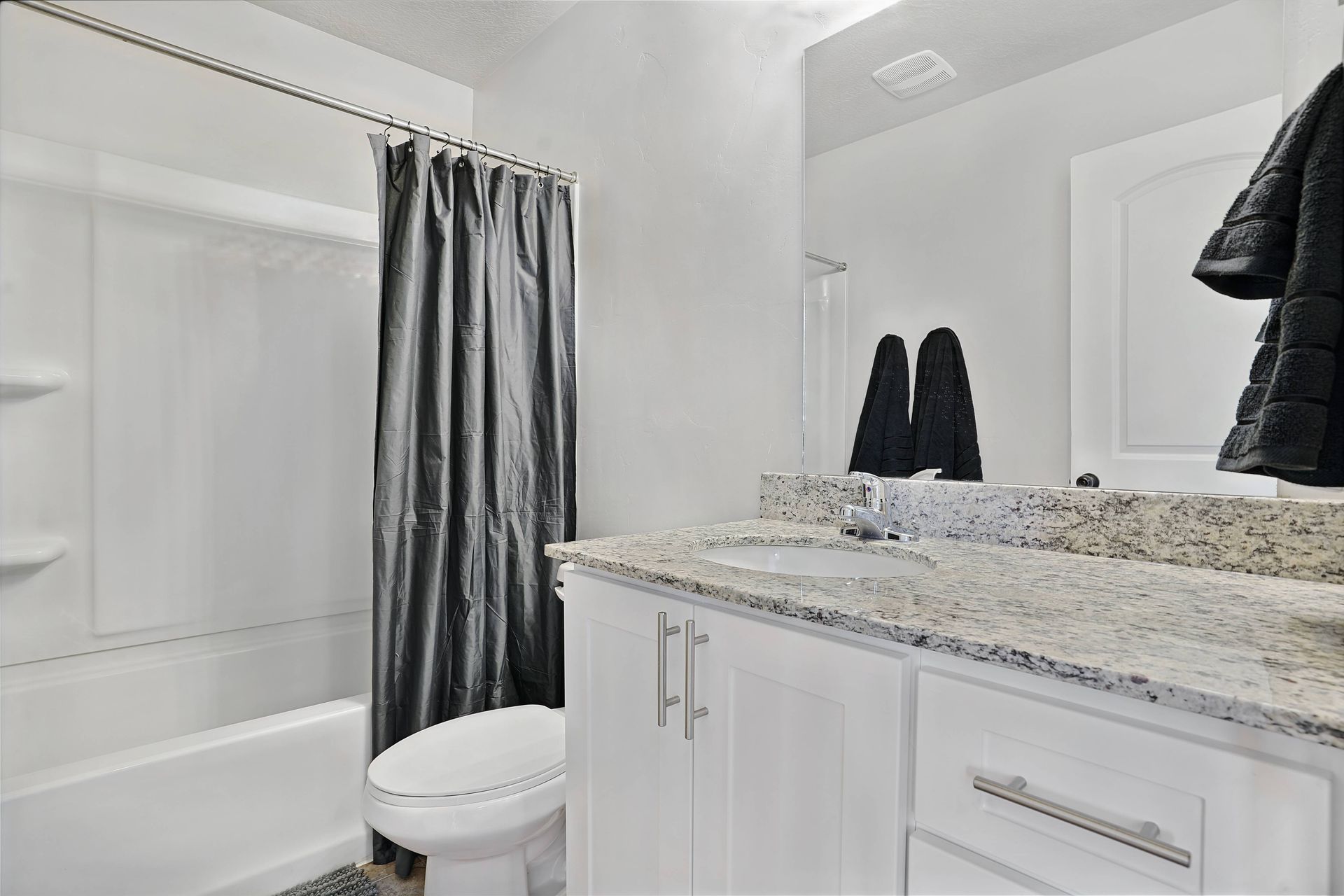 Bathroom with white cabinets, speckled countertop, toilet, bathtub with gray curtain, and mirror with towels.