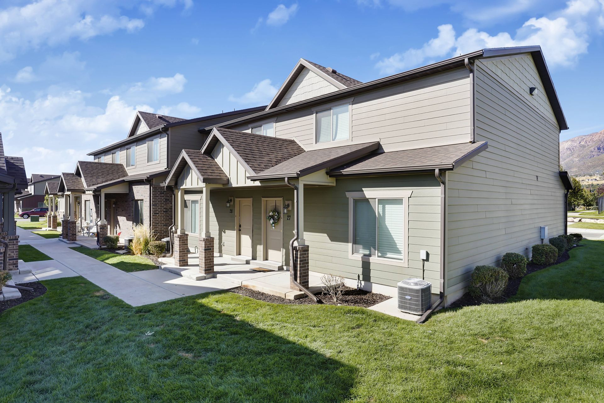 Townhomes with green siding and brown roofs, set in a grassy area with a mountain backdrop.