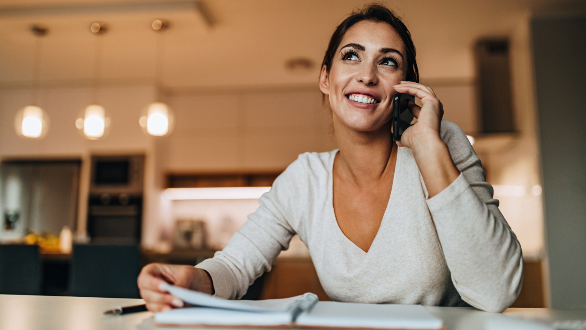Woman smiling on phone at a kitchen table, writing in a notebook under warm pendant lights