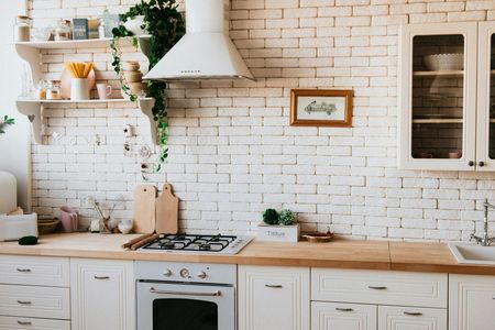 White-painted brick kitchen with wooden countertops, stove, and shelves, decorated with plants and cooking items.