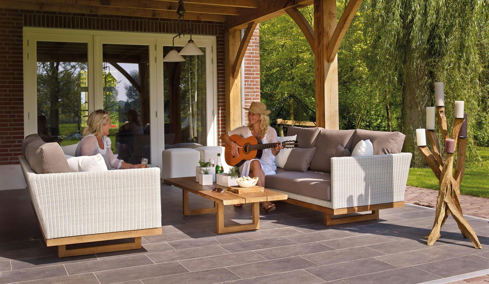 Two women relax on patio furniture, one playing guitar. Sunny setting.