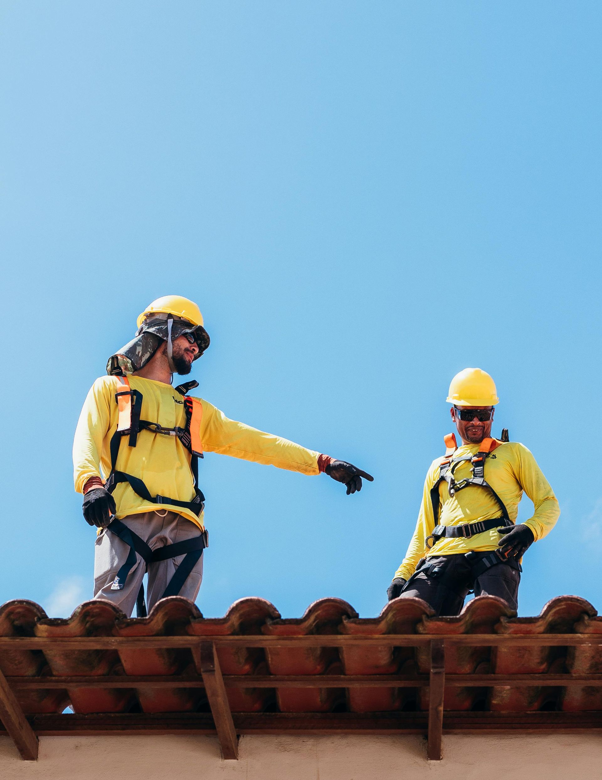 Two roofers in yellow safety gear on a tile roof point to something, blue sky background.