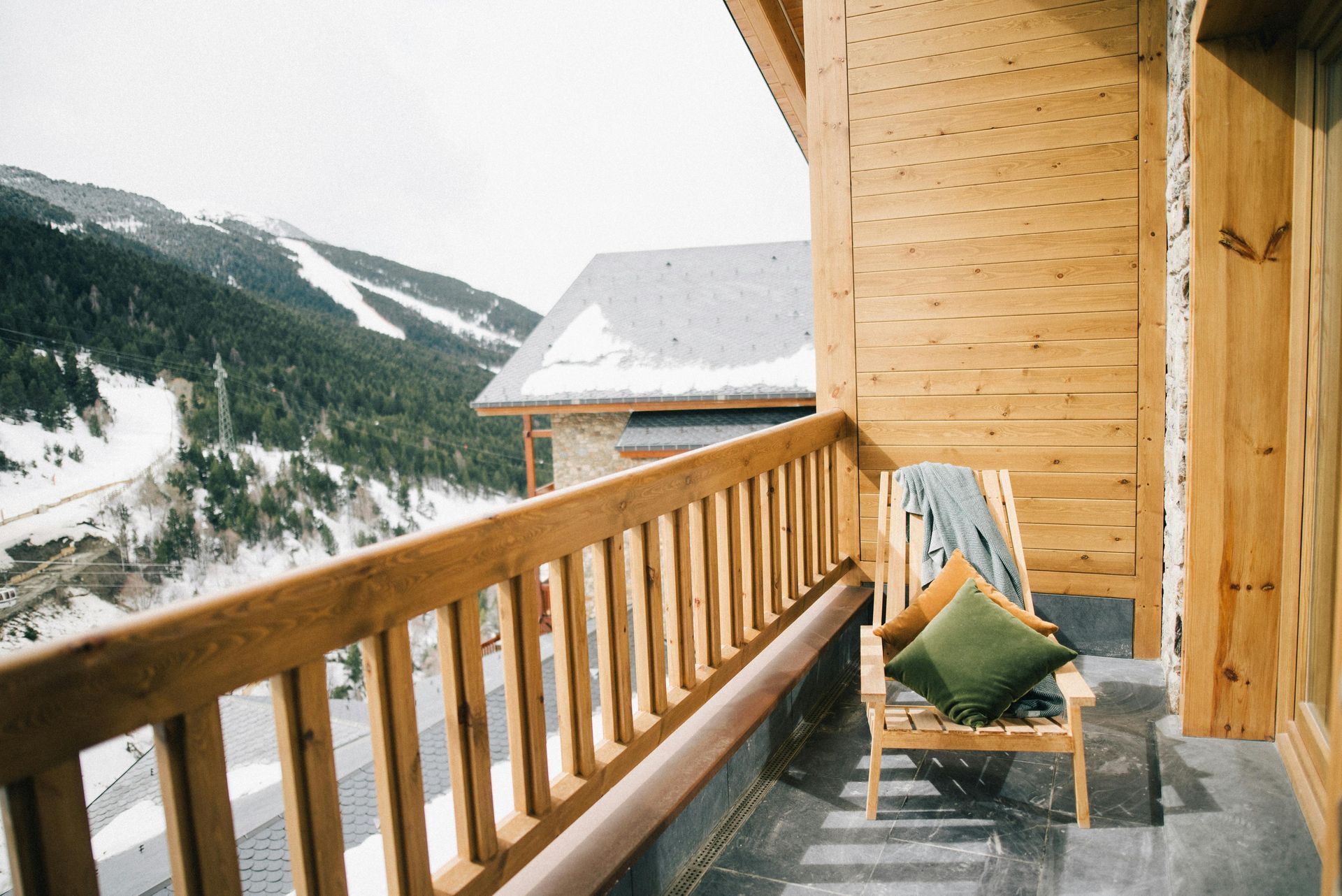 Wooden balcony overlooking snowy mountains; chair with pillows and blanket.