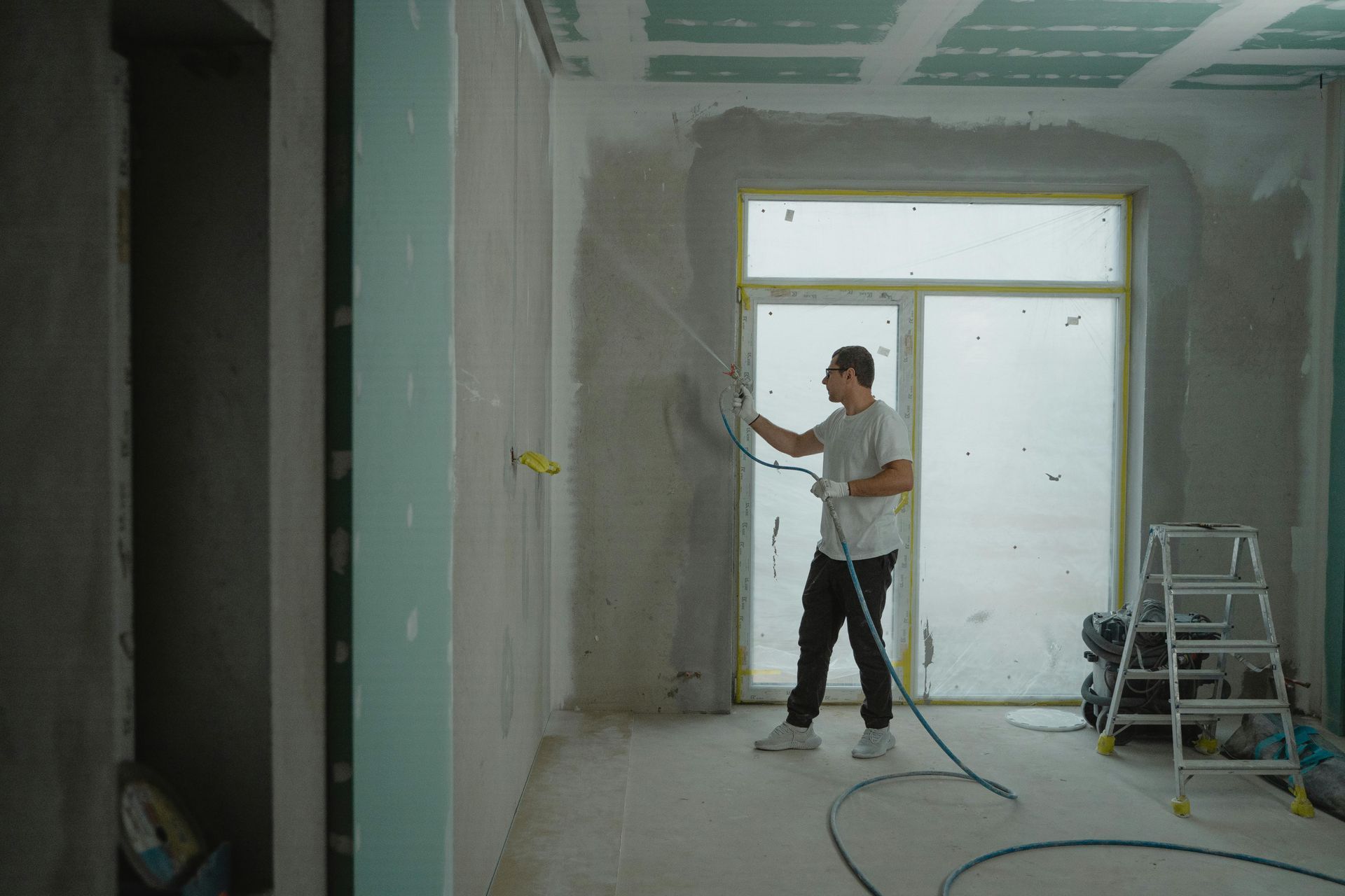 Man sprays plaster on a wall in a room during renovation. He is holding a sprayer and wearing white shirt and black pants.