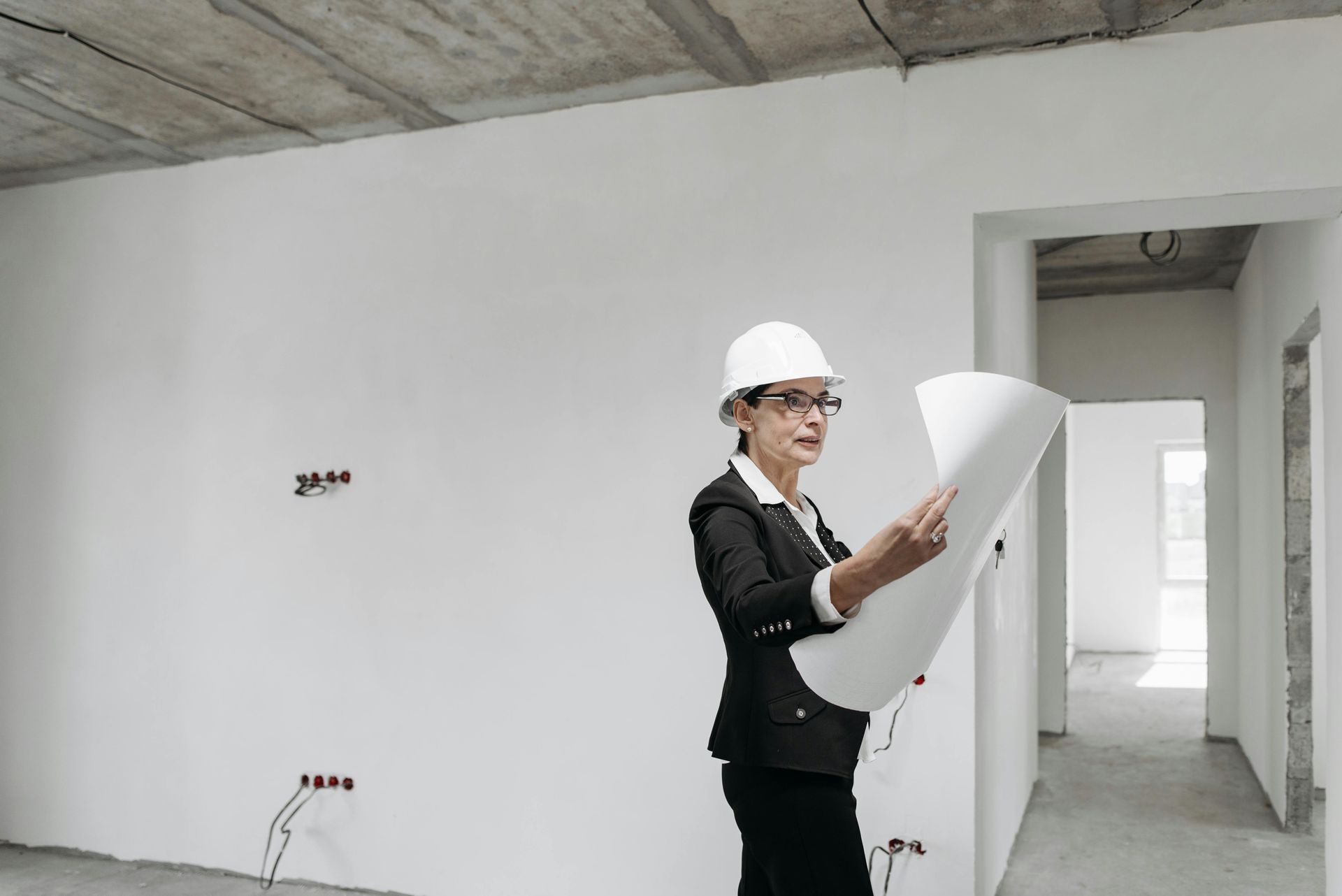 Woman in hard hat, blazer, looking at blueprints inside a construction site.