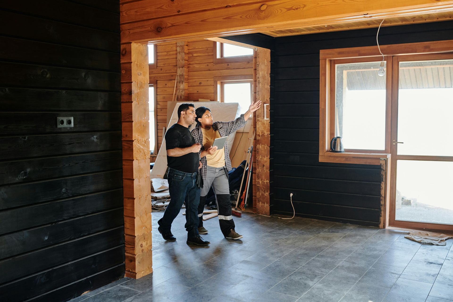 Two people in a room under construction, one pointing upwards. Black walls, wooden beams, and large windows.