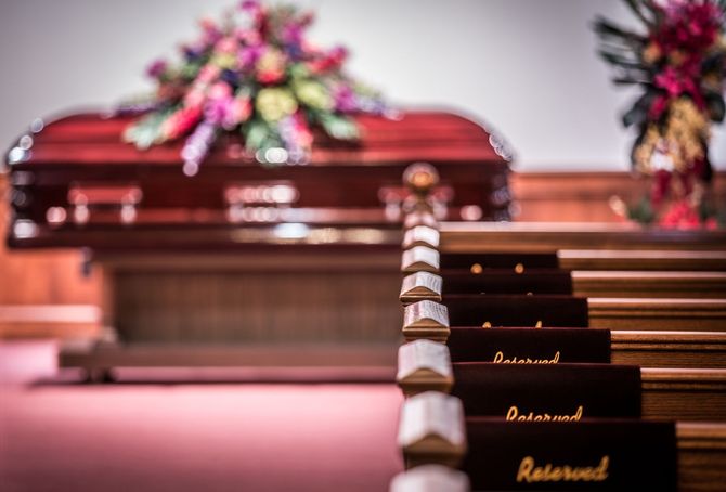 Open casket at a funeral with pink flowers and rows of dark chairs in the chapel