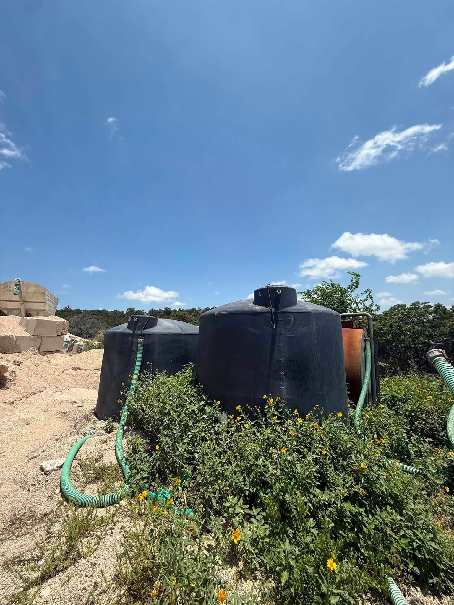 Black water tanks and a green hose amid overgrown plants under a bright blue sky