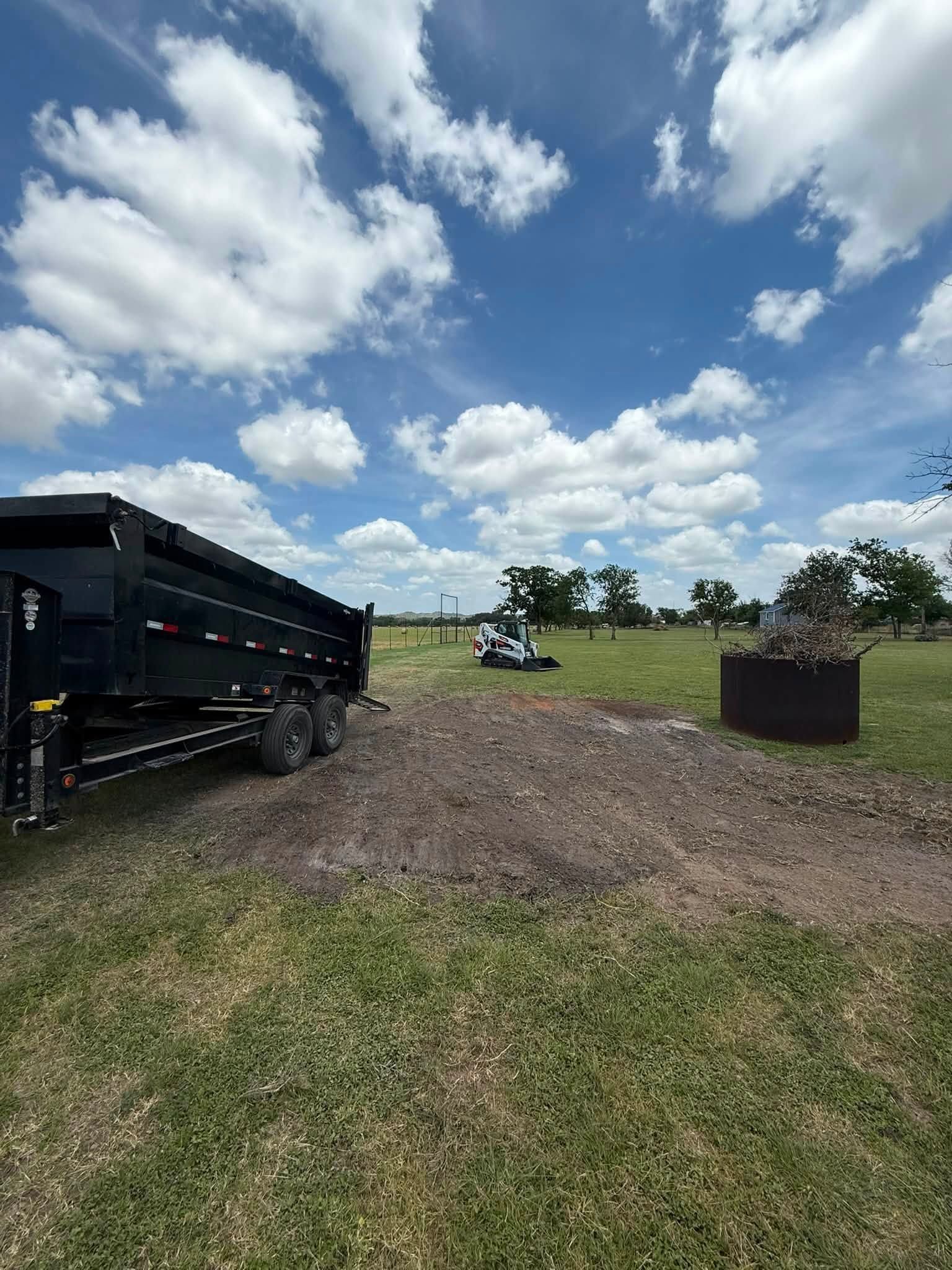 Black dump trailer on gravel field under a cloudy blue sky, with a burn barrel and tractor in the distance