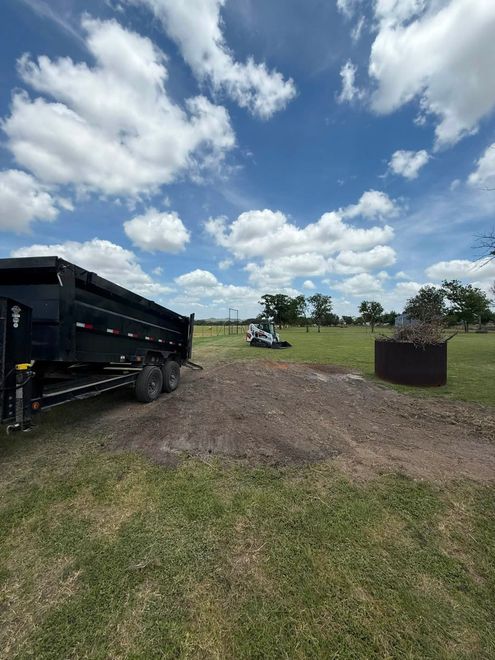 Black dump trailer on gravel field under a cloudy blue sky, with a burn barrel and tractor in the distance
