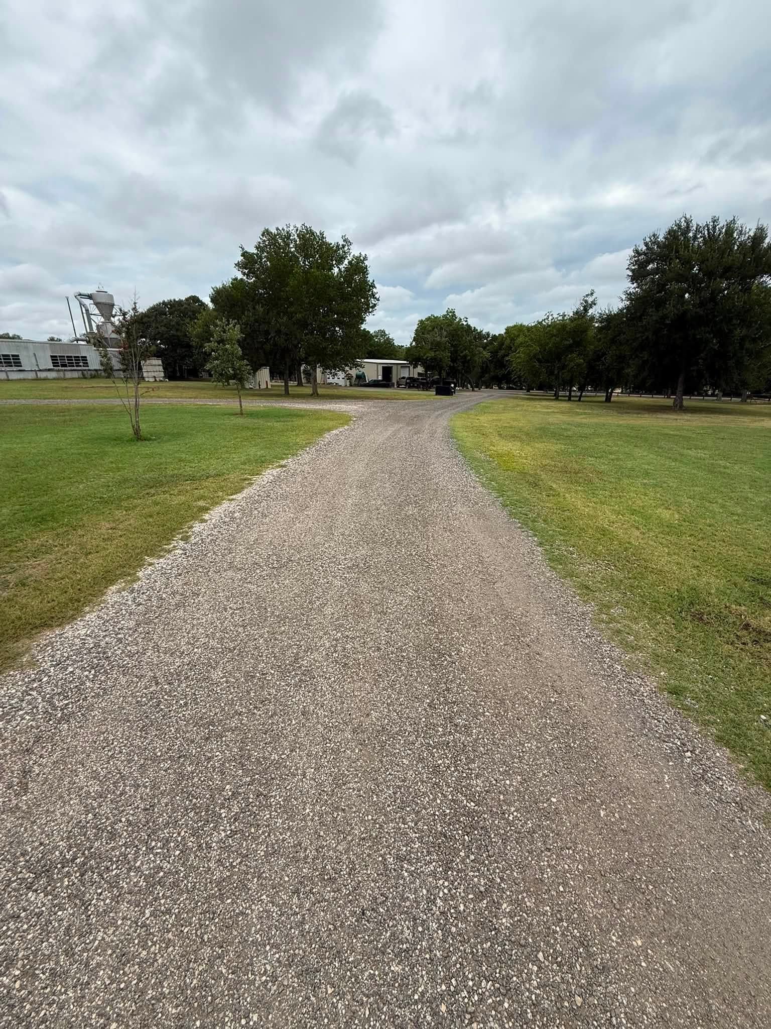 Gravel path through a grassy park leading to trees and houses under a cloudy sky