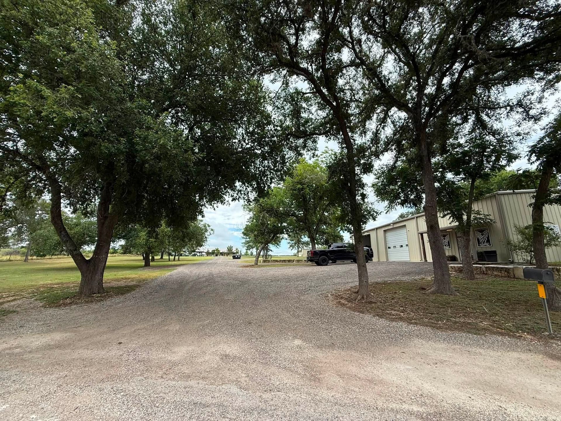 Gravel driveway lined with trees leading to a parked car and small white building on the right
