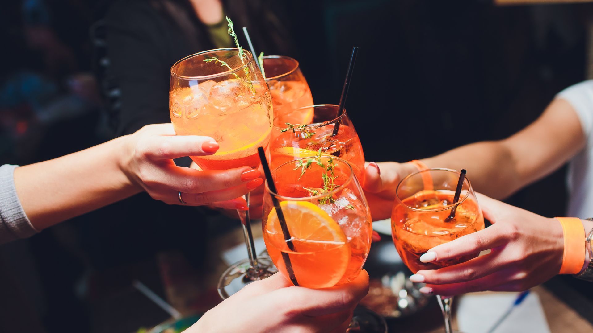 Menschen stoßen feierlich mit schön hergerichteten Cocktailsgläsern an, darin befindet sich ein orangenes Getränk mit Orangen.