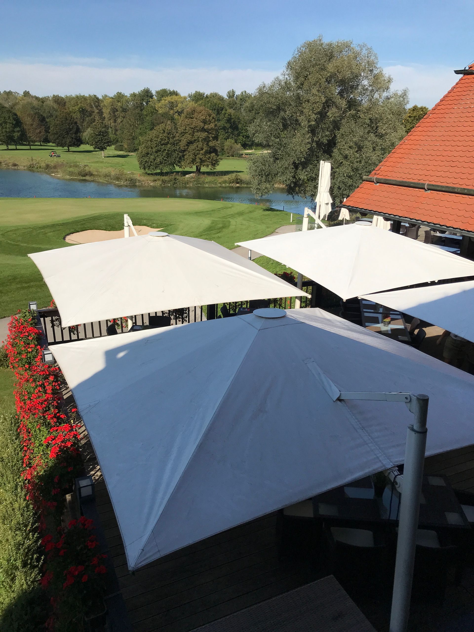 Blick von oben auf die Sonnenschirme der Terrasse des Restaurant Platzhirsch, daneben sieht man Blumen und den Golfplatz