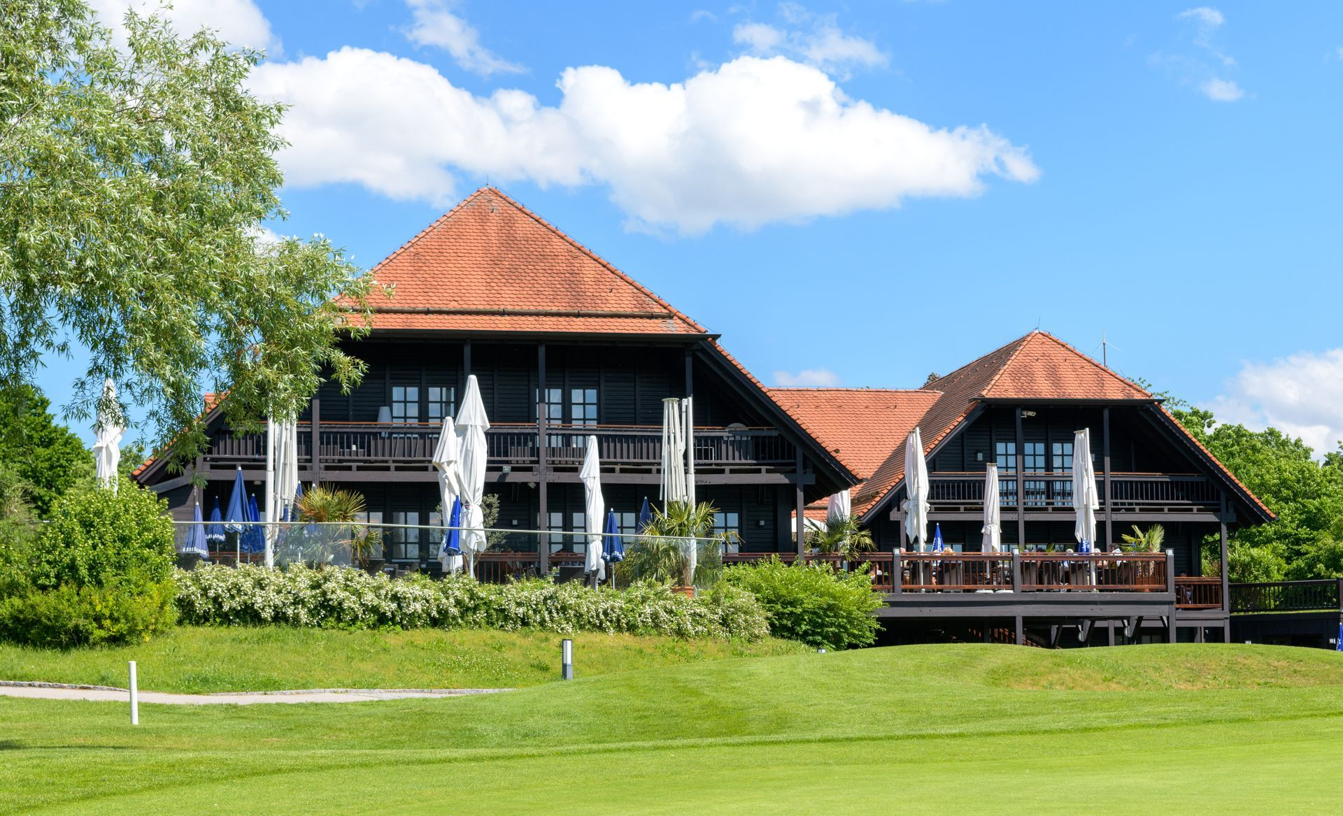 Blick auf die Terrasse des Restaurant Platzhirsch vom Golfplatz aus. Man sieht das Gebäude und Sonnenschirme