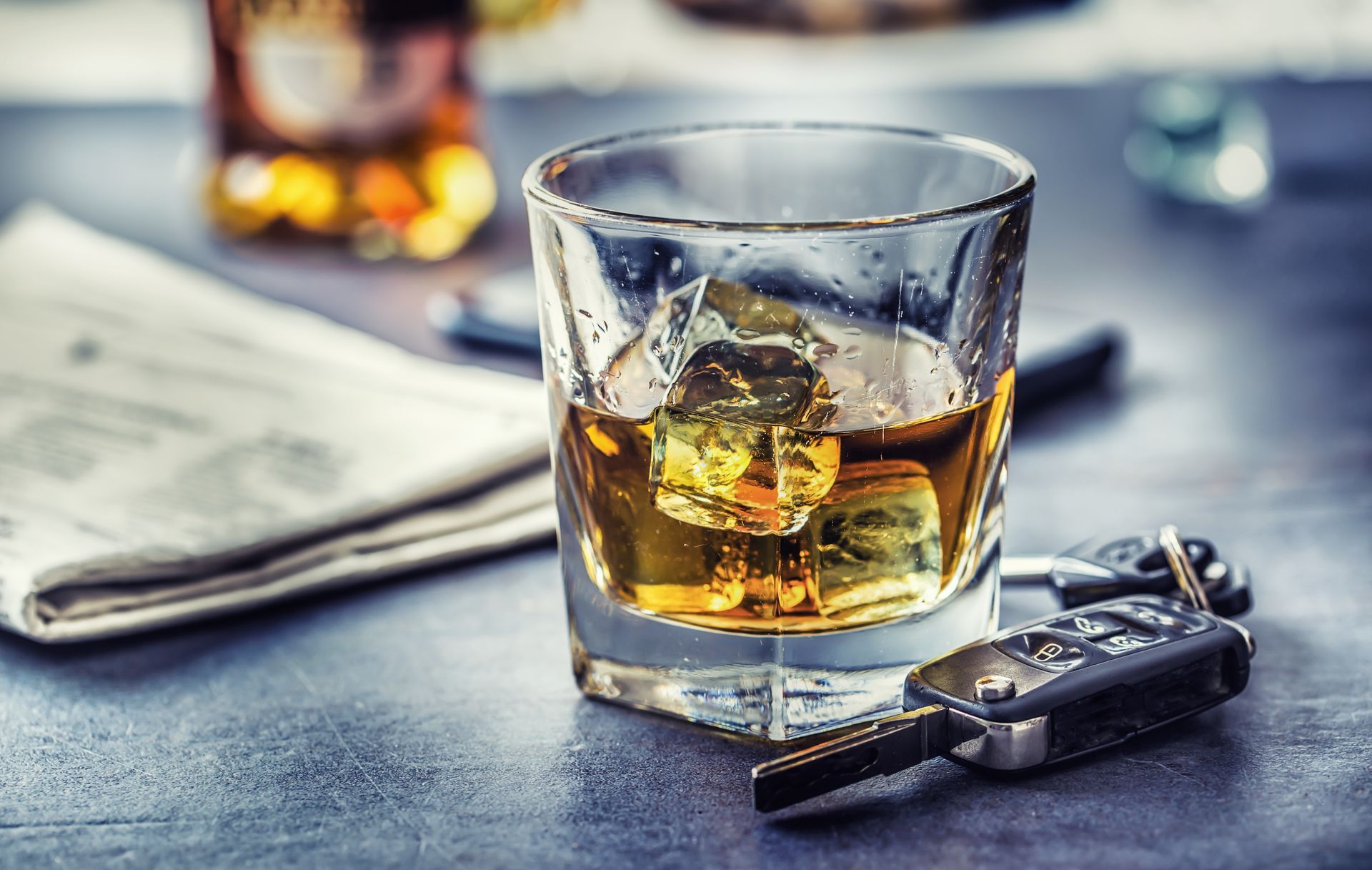 Photograph of a glass containing whiskey with ice cubes placed on a dark surface next to a set of car keys and a folded newspaper in the background. Photograph of a glass containing whiskey with ice cubes placed on a dark surface next to a set of car keys and a folded newspaper in the background.