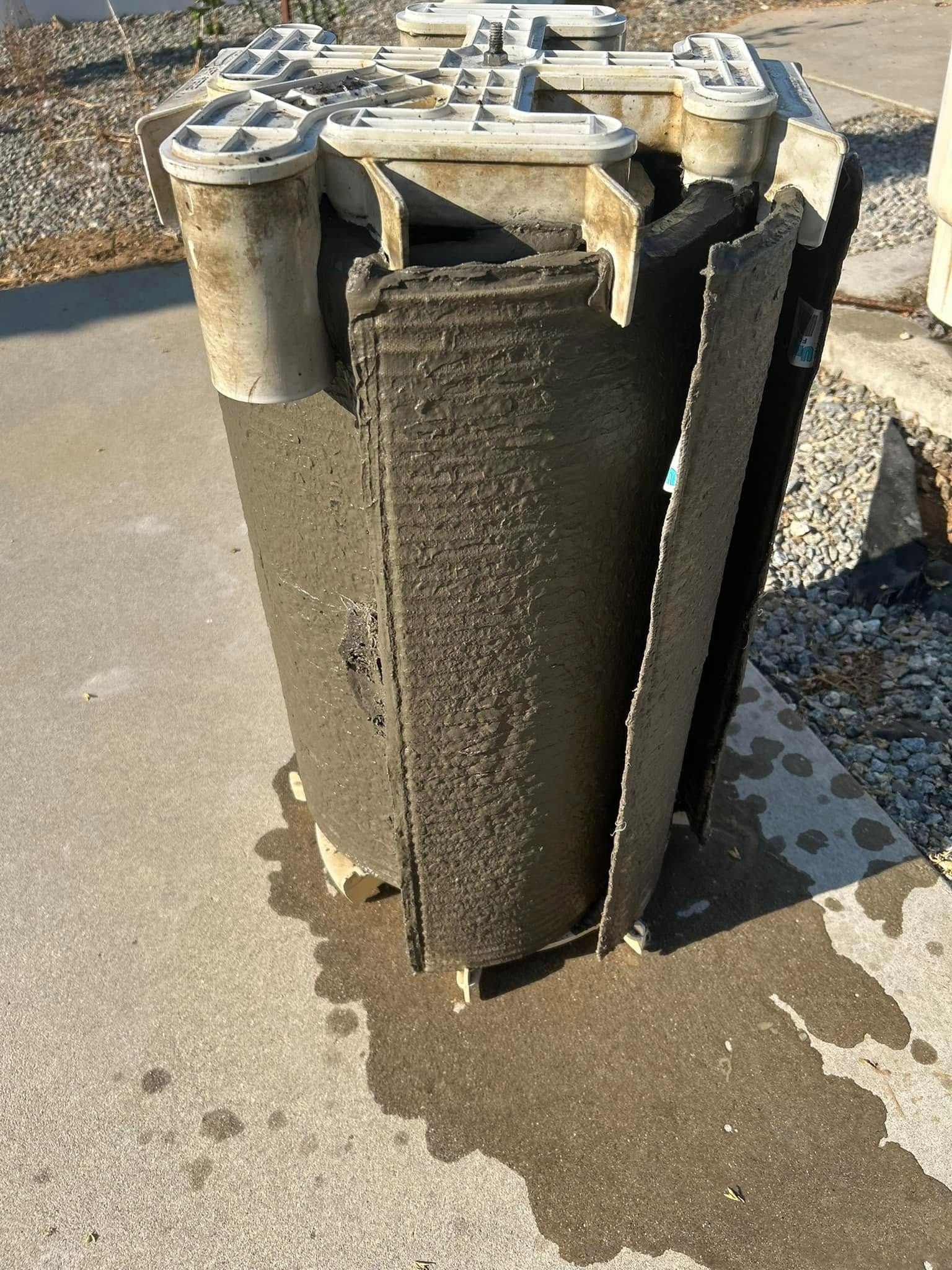 A cylindrical water filter, partially disassembled, sits on concrete. It is dirty, and water has dripped onto the surface.