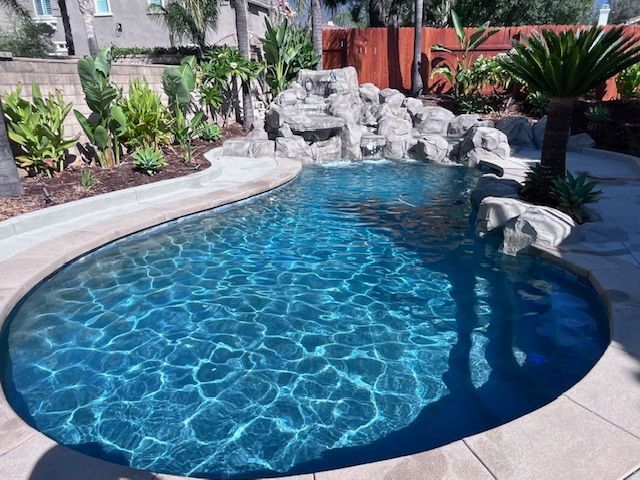 Swimming pool with waterfall, surrounded by plants and stone, in a backyard setting.