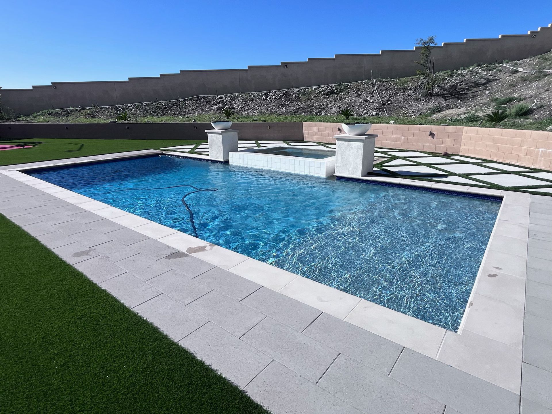 Rectangular pool with blue water and stone patio, surrounded by artificial turf and a retaining wall.