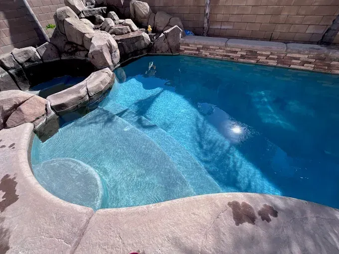 Pool with waterfall feature and built-in steps, filled with clear blue water, surrounded by concrete.