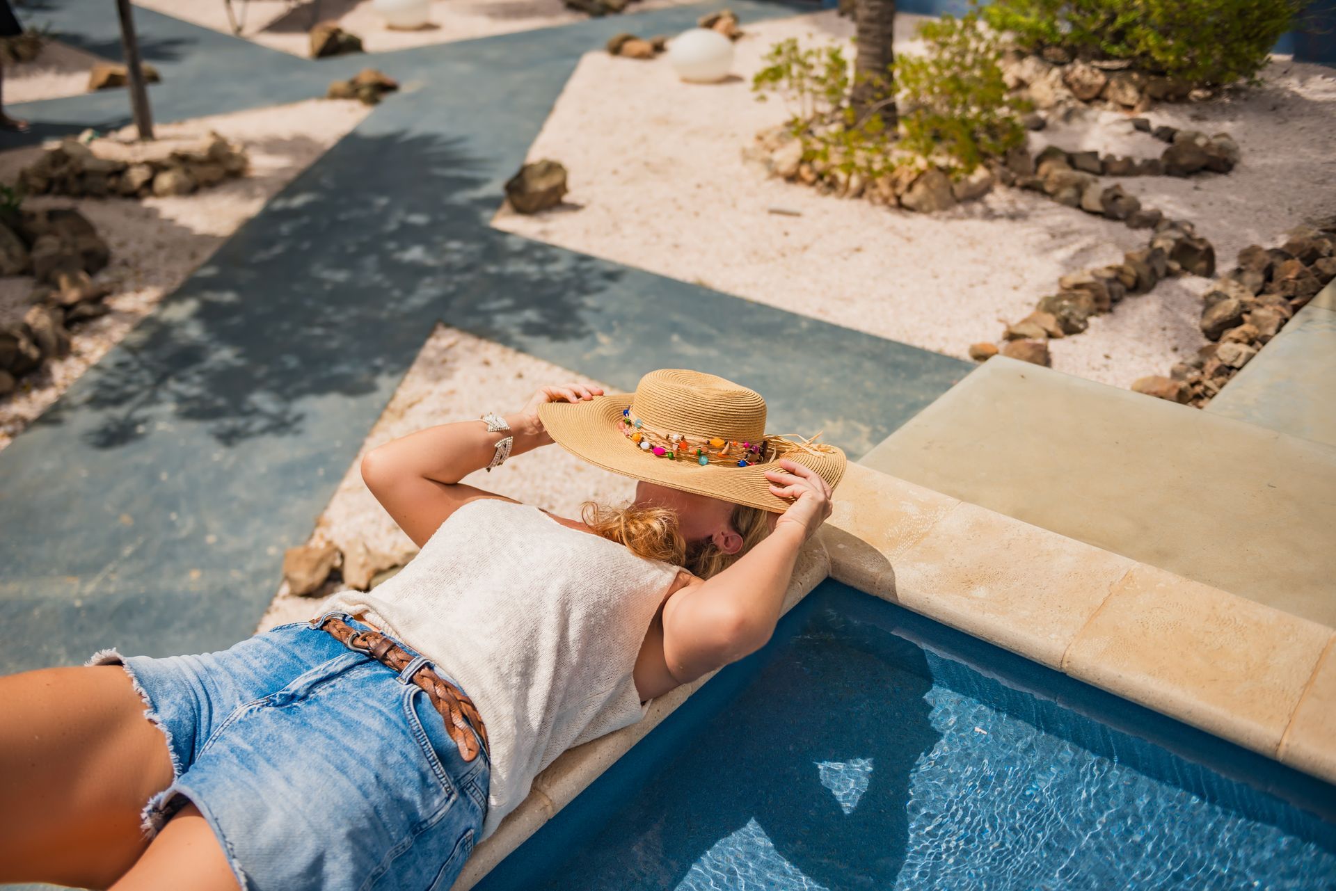 A woman is laying on the edge of a swimming pool wearing a straw hat.