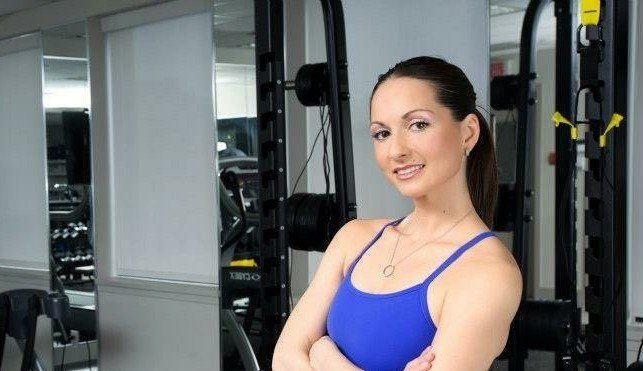 A woman in a blue tank top is standing in a gym with her arms crossed.