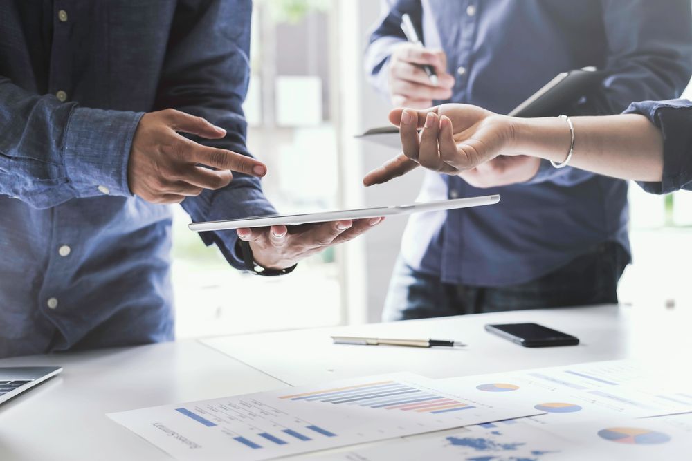 People discussing work, looking at charts, and using a tablet in an office setting.