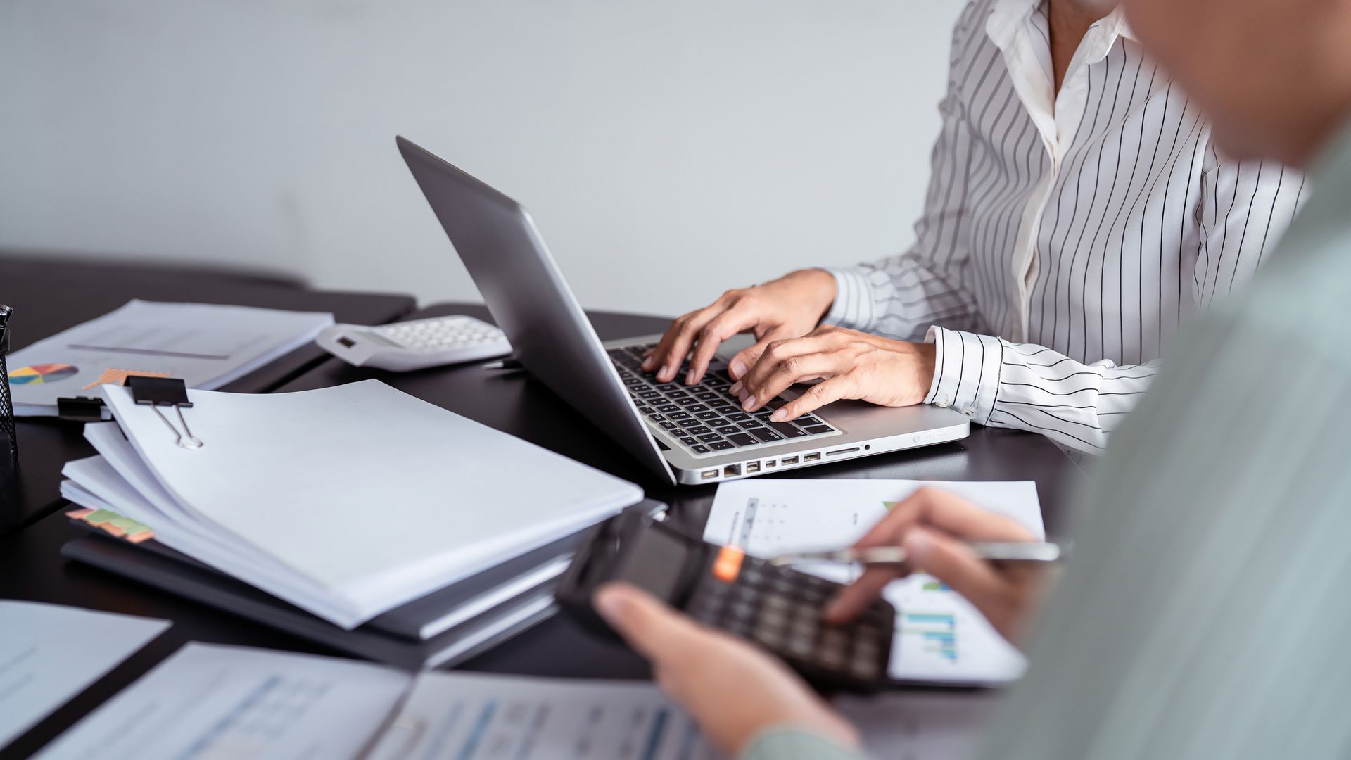 Two people working at a desk with a laptop and calculator, surrounded by stacks of paper documents.