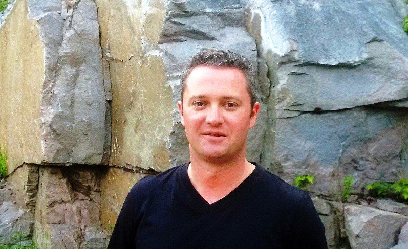 A man in a black shirt is standing in front of a rock wall.