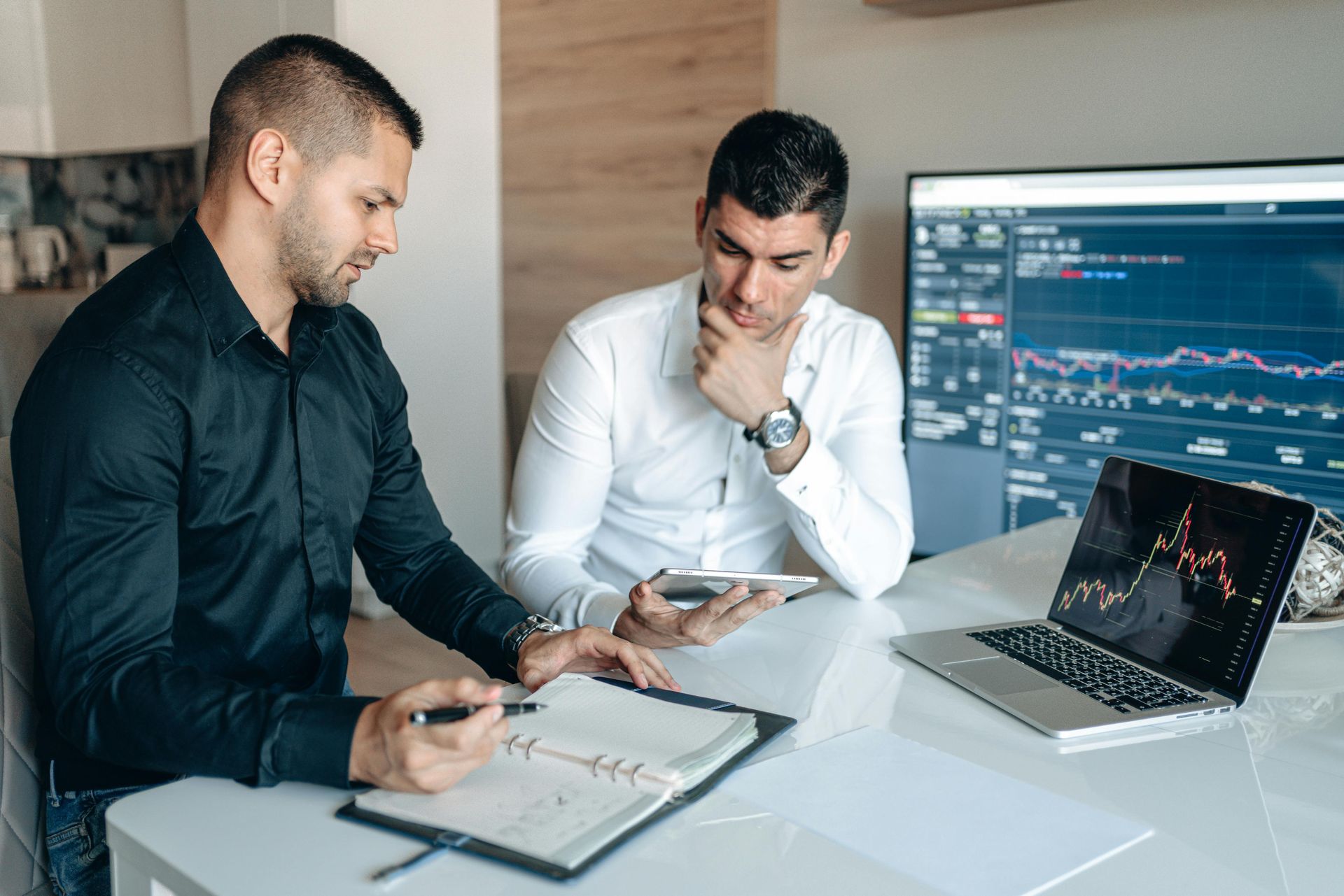 Two men analyzing financial data, one pointing at a notebook, the other holding a tablet, with a computer screen and laptop displaying charts.