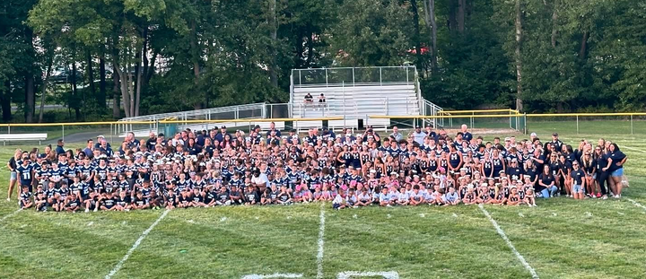 A large group of people on a football field. They are posing in front of bleachers and trees.