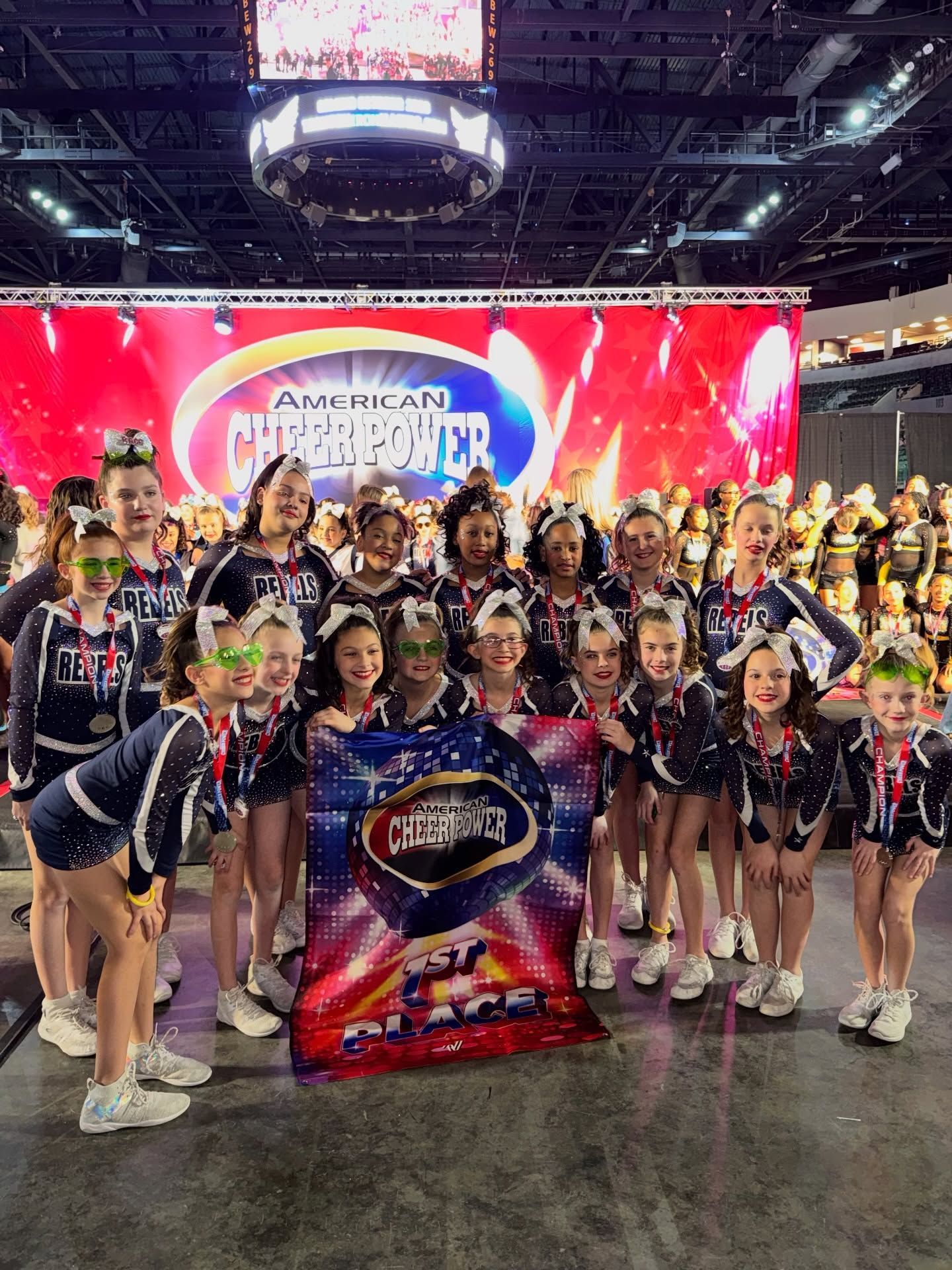 Cheerleading team posing with championship banner, wearing blue and white uniforms, in arena.