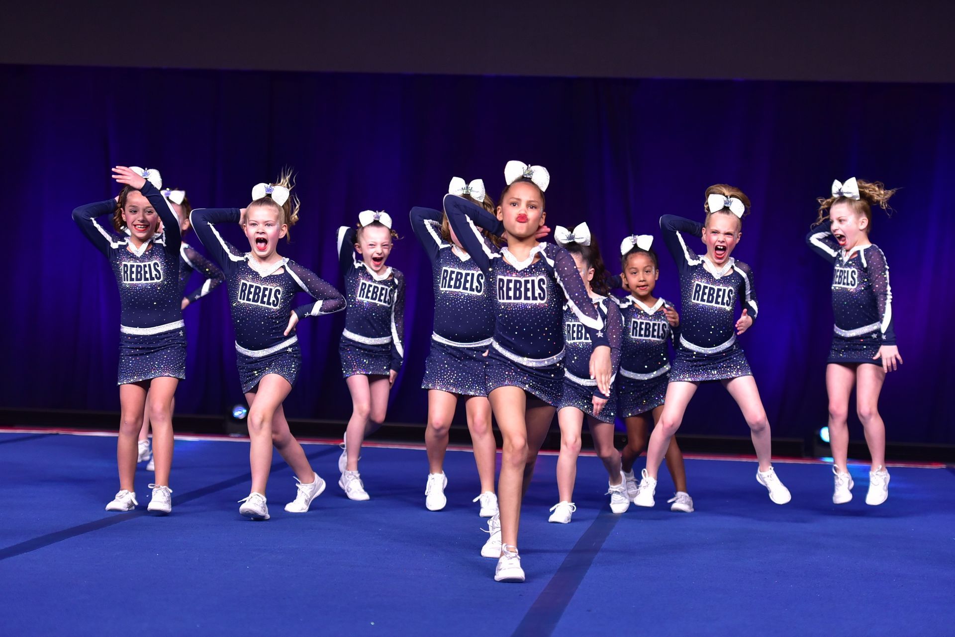 Cheerleaders in blue uniforms with white bows perform a routine on a blue mat, raising arms and shouting.