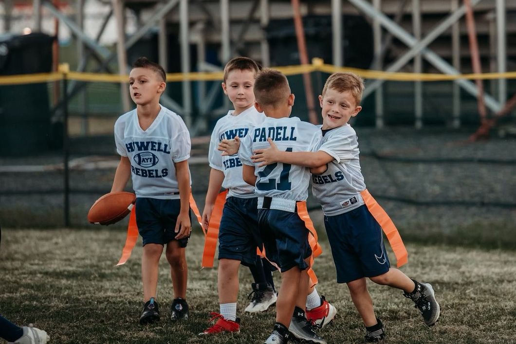 Youth flag football game. Four boys in white jerseys and blue shorts on a green field. Two boys hug, one with a football.