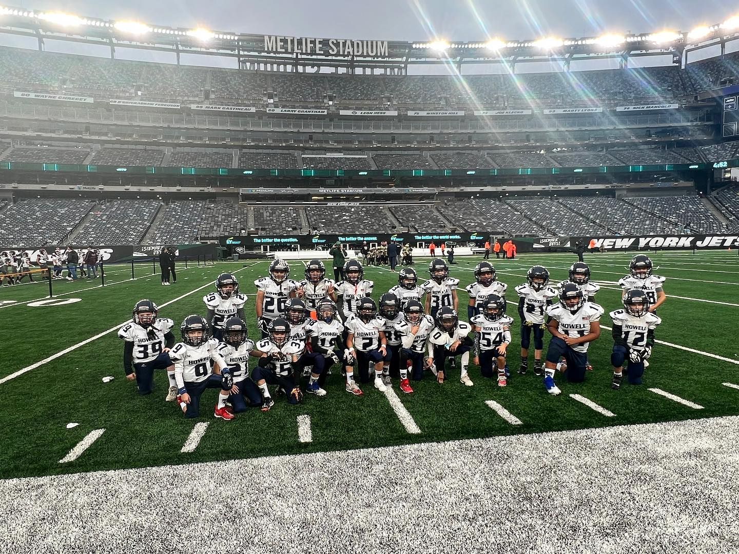 Youth football team on a green field in a large stadium. Players in uniforms, kneeling, with stadium seating in background.