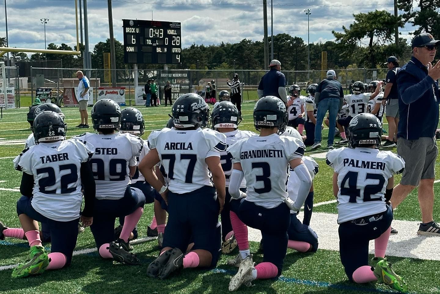 Football team kneeling on the field. White jerseys, navy helmets, pink socks. Coach stands nearby. Scoreboard in background.