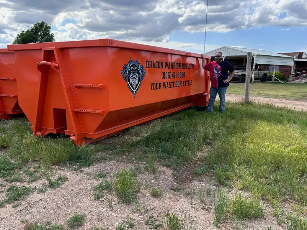 A black dumpster is sitting in the grass next to a tree.