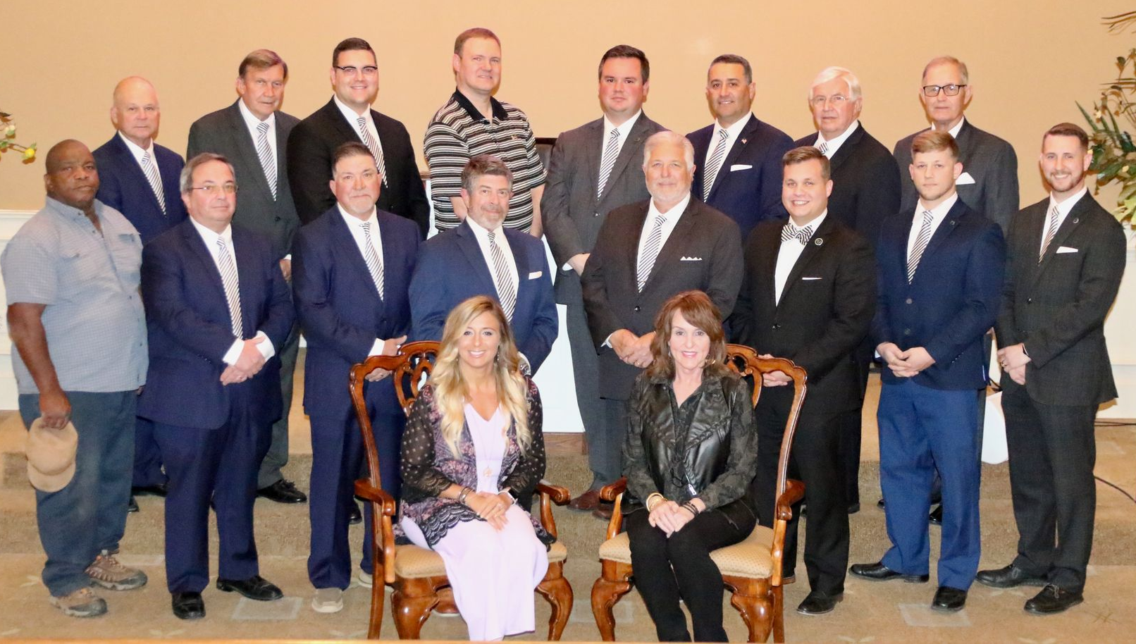 Group of men and two women posing for a photo; most are wearing suits, while two are casually dressed. They are indoors.