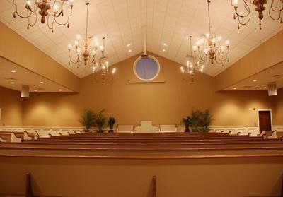 Interior of a chapel with tan walls, rows of pews, and chandeliers. A round window is centered above a small stage.