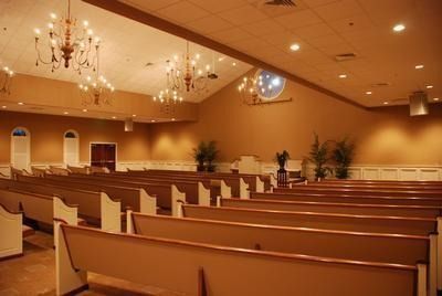 Interior of a church with rows of wooden pews facing a raised platform. Tan walls, ornate chandeliers, and a circular window are visible.